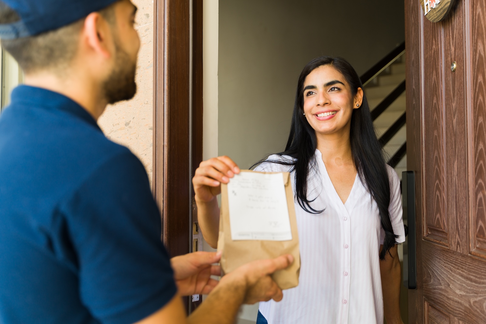 Cheerful young woman is receiving a package with medicine from a delivery man at the door of her home