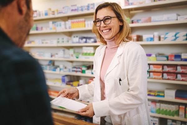 Smiling female pharmacist with prescription assisting a customer standing at the counter. Helpful female pharmacist dealing with a male customer standing at her desk in the pharmacy, she is smiling as she explains a prescription.